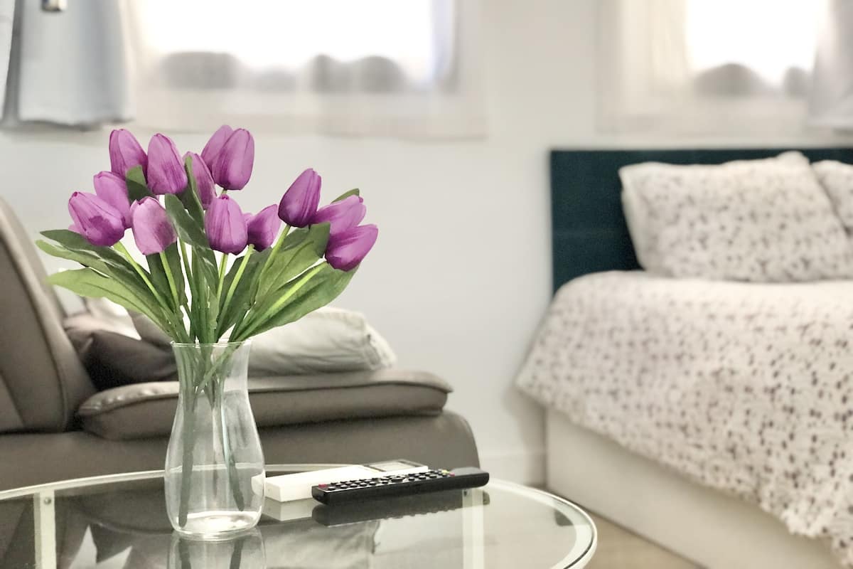 A clear glass vase with purple flowers on a glass table in a room with a bed and a remote control.