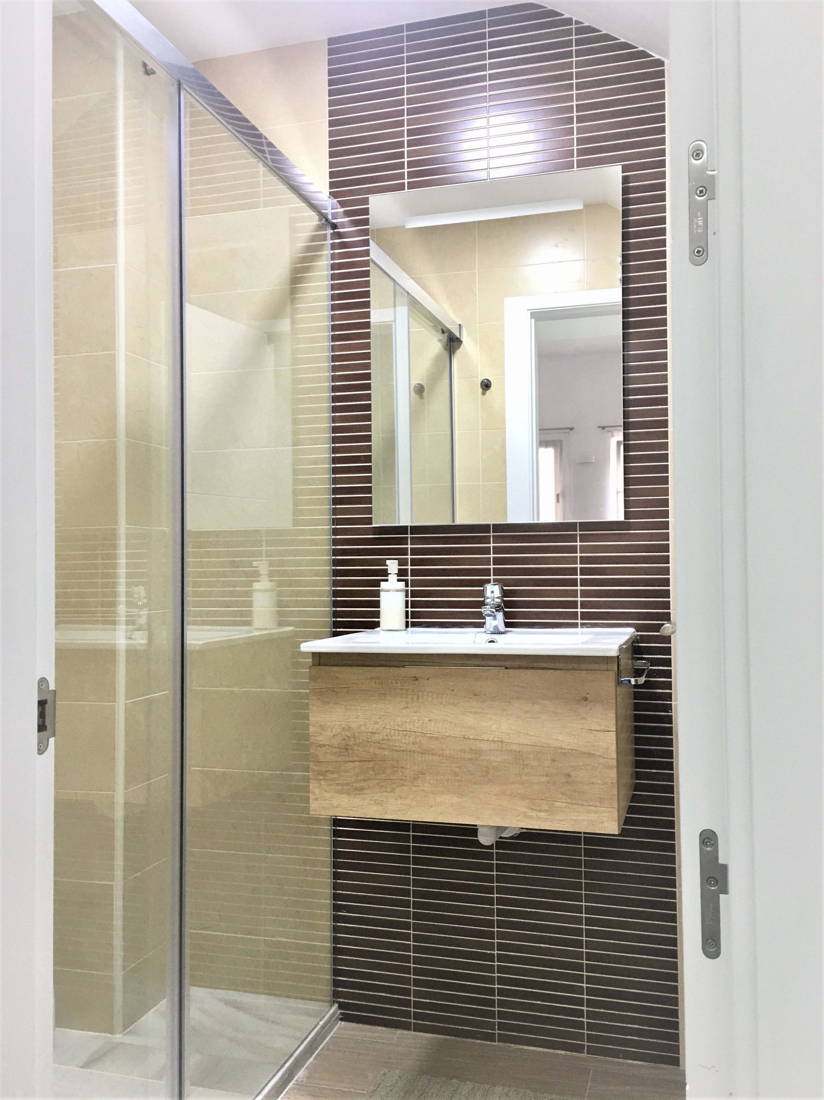 Une salle de bain dotée d’une douche à parois de verre, d’un meuble-lavabo en bois avec un miroir et d’un pare-feu en carreaux.