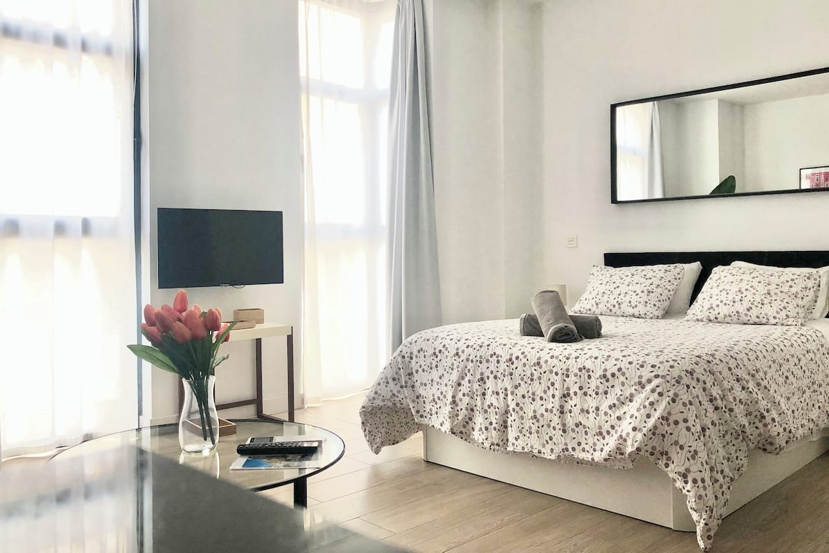 A hotel room with a bed, a TV, a small table with a vase of flowers, and a window with curtains.