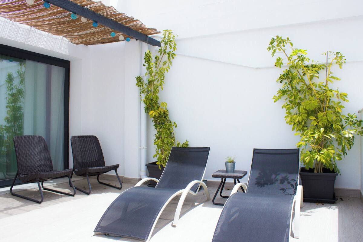 A sunlit outdoor seating area with two lounge chairs and a small table, surrounded by potted plants and a pergola.