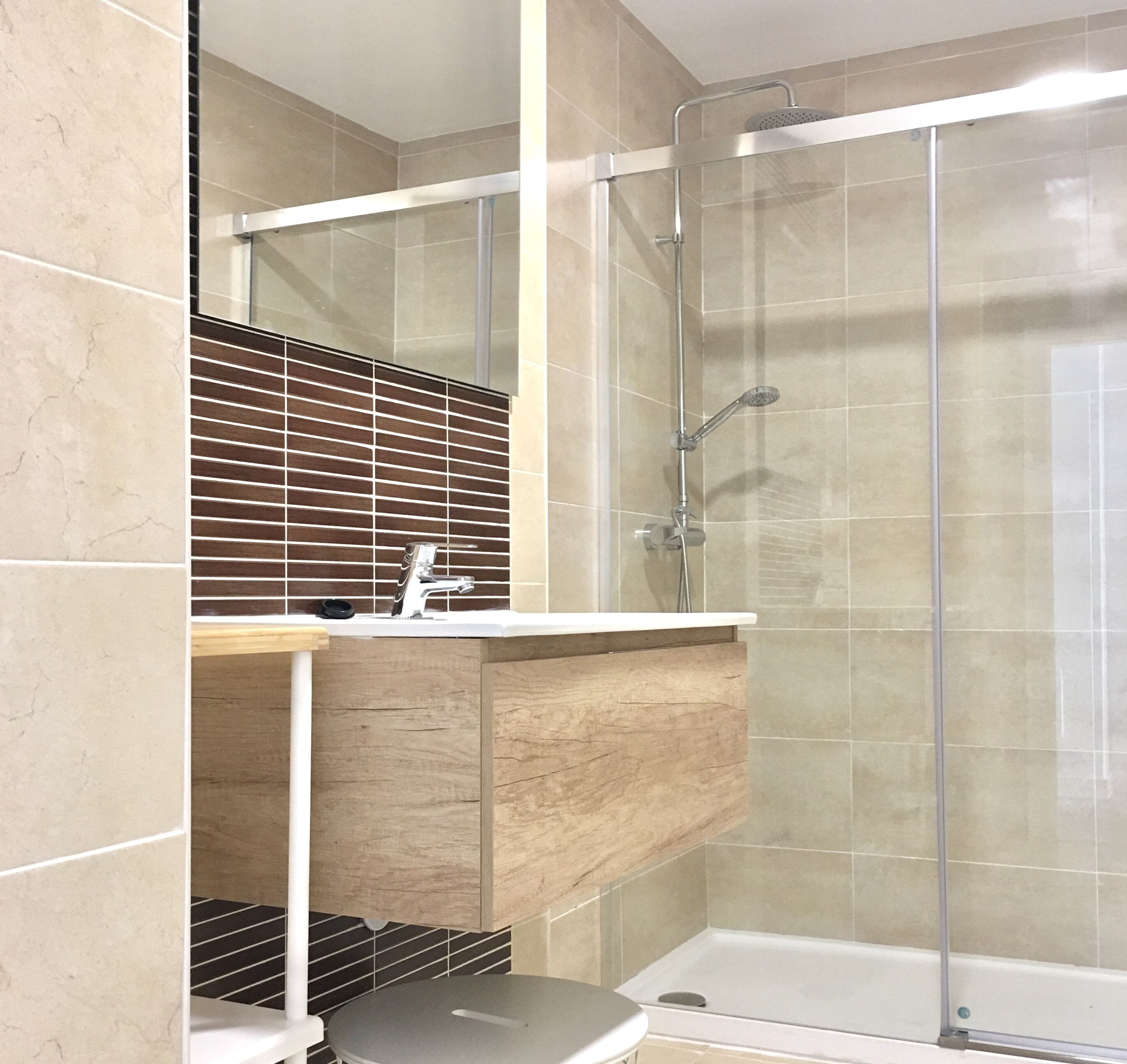 Un baño moderno con ducha de vidrio, un lavamanos con espejo y una mesita pequeña.