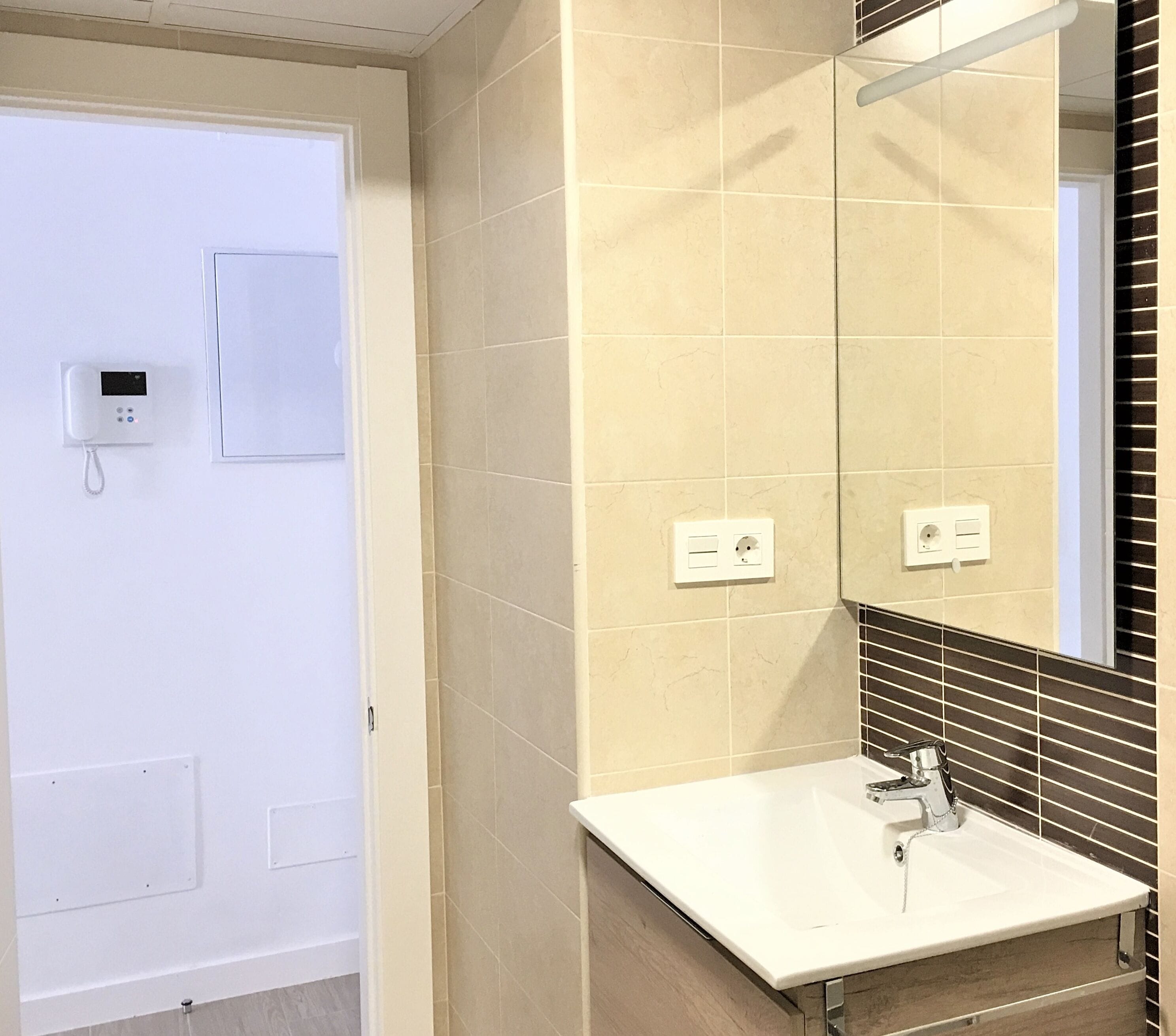 A bathroom with beige tiles, a white sink, a mirror, and a door with a phone and light switch.