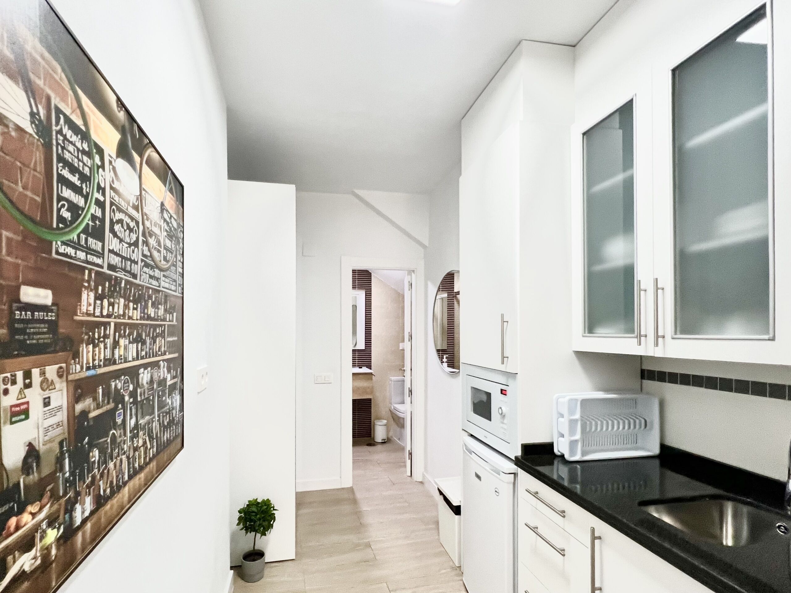 A narrow kitchen with white cabinets, a black countertop, and a microwave. There is a small potted plant and a bar area with liquor bottles on the left.