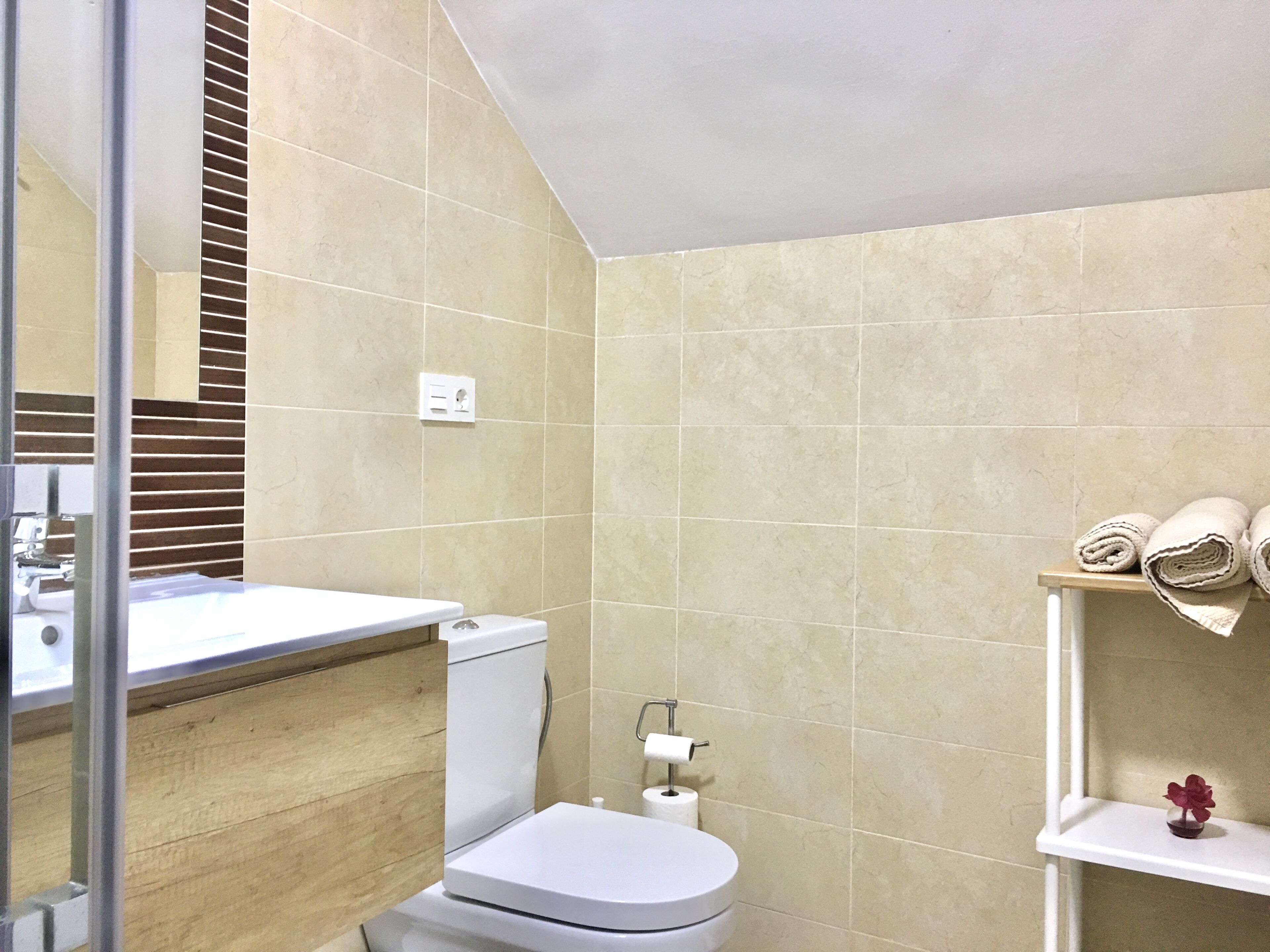 A bathroom with beige tiles, a white toilet, a sink with a mirror, and a shelf with towels.