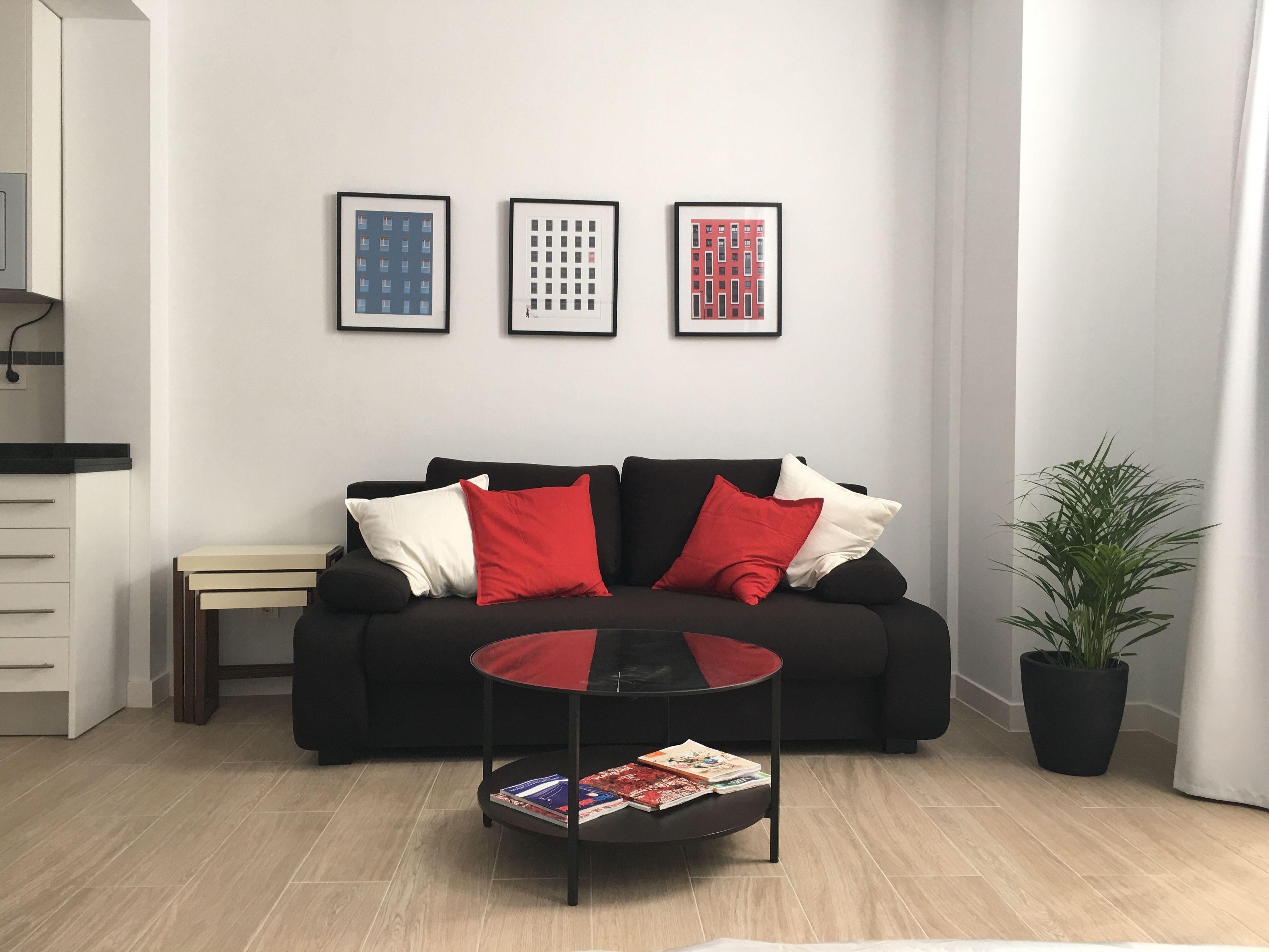 En moderne stue med en sort sofa, røde og hvide puder, et glasbord med magasiner og tre billeder i rammer på væggen.