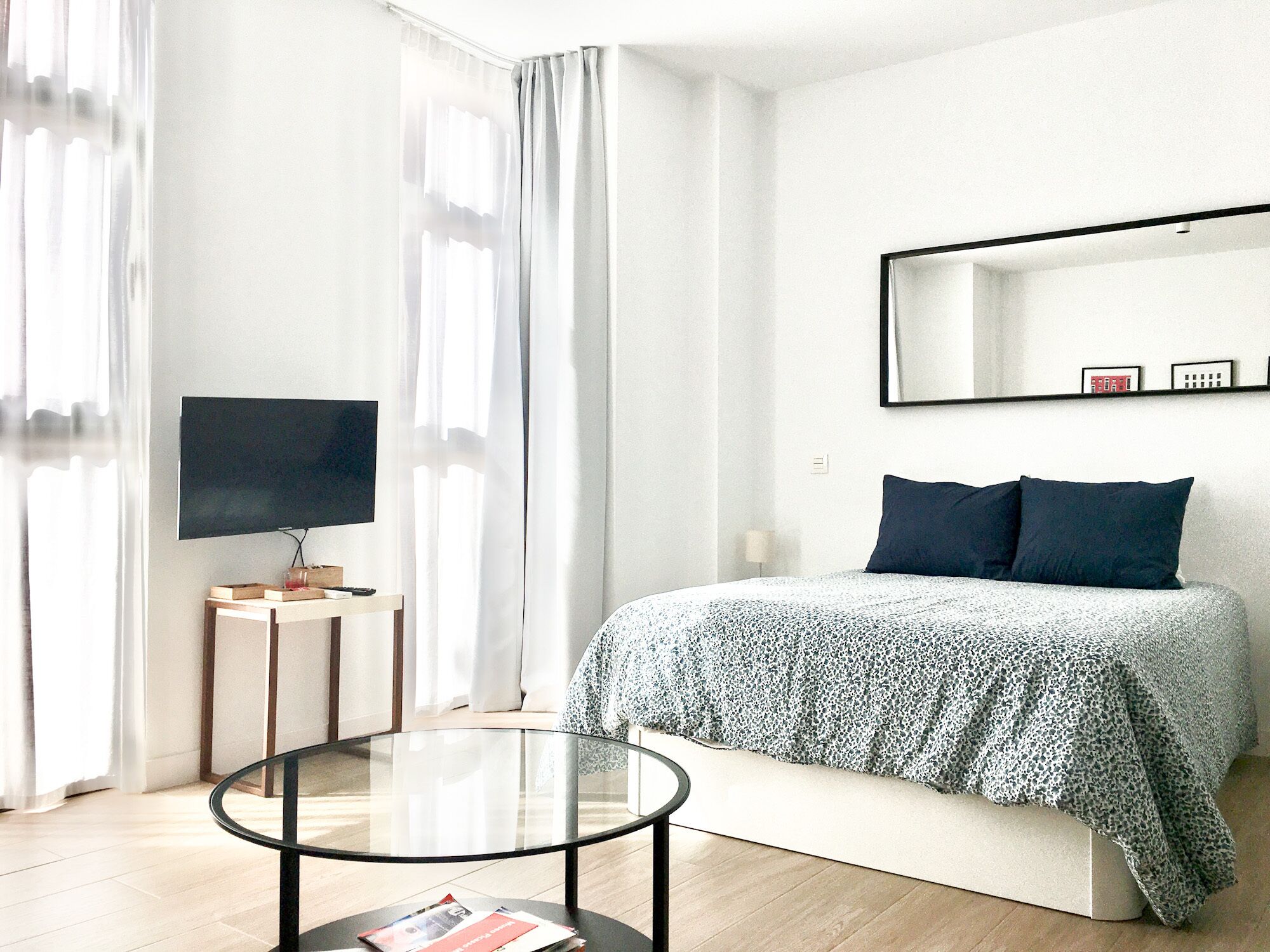 A modern bedroom with a bed, a TV, a small table, and a mirror on the wall.