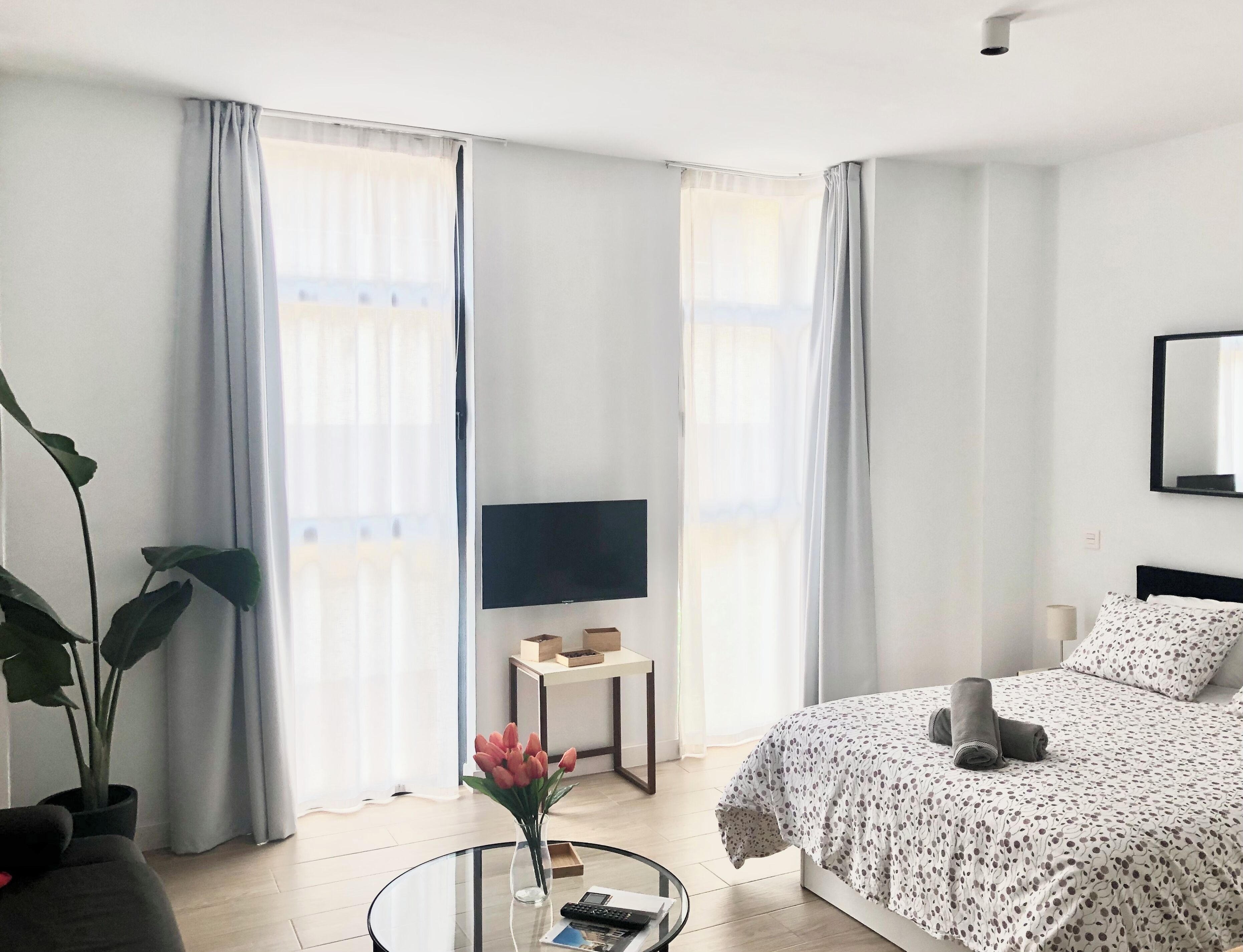 Een moderne hotelkamer met een bed, een televisie, een klein tafeltje met een bloevas en een groot raam met gordijnen.