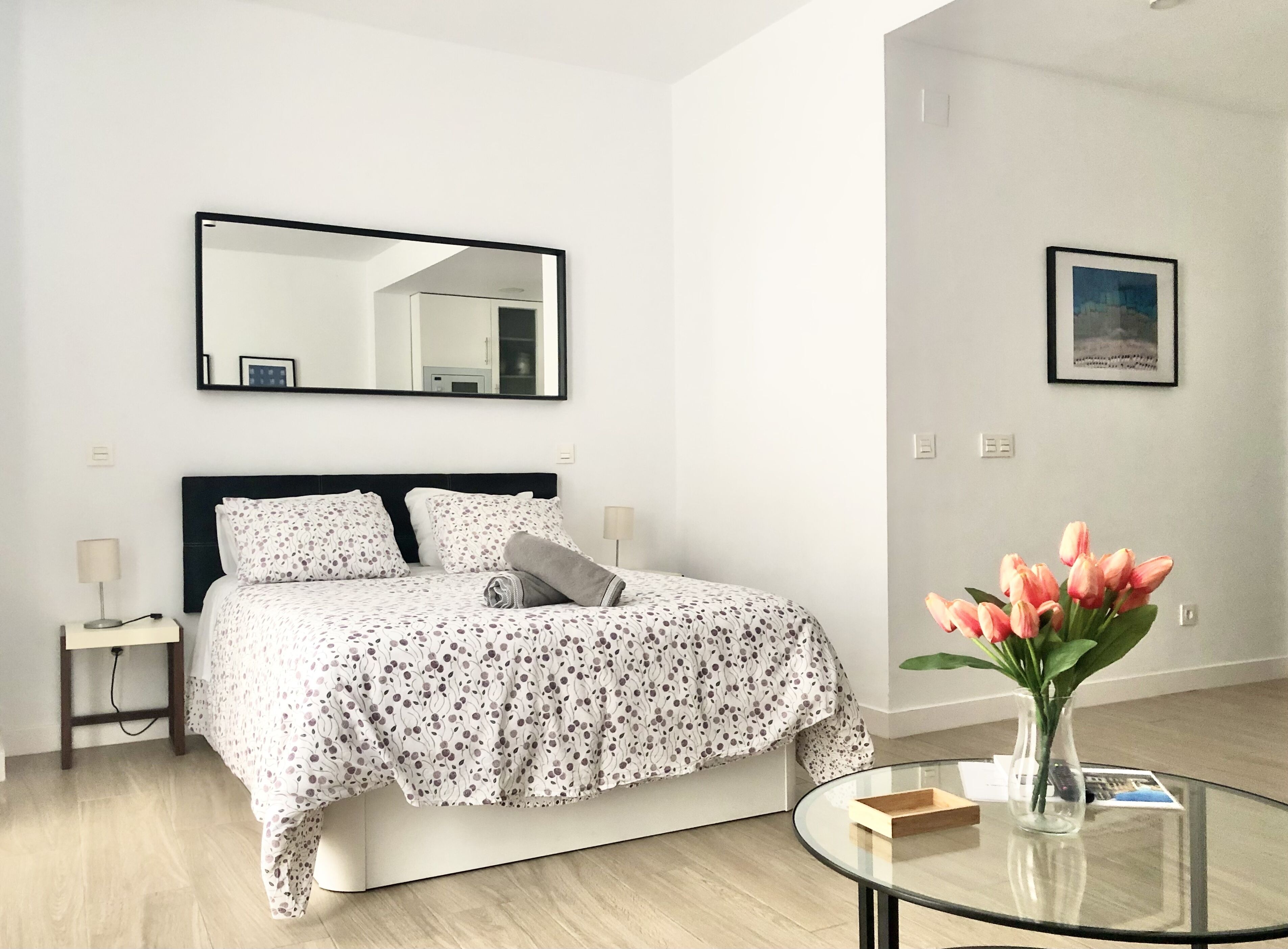 A modern bedroom with a bed, a bedside table, a mirror, a glass coffee table, and a vase with flowers.