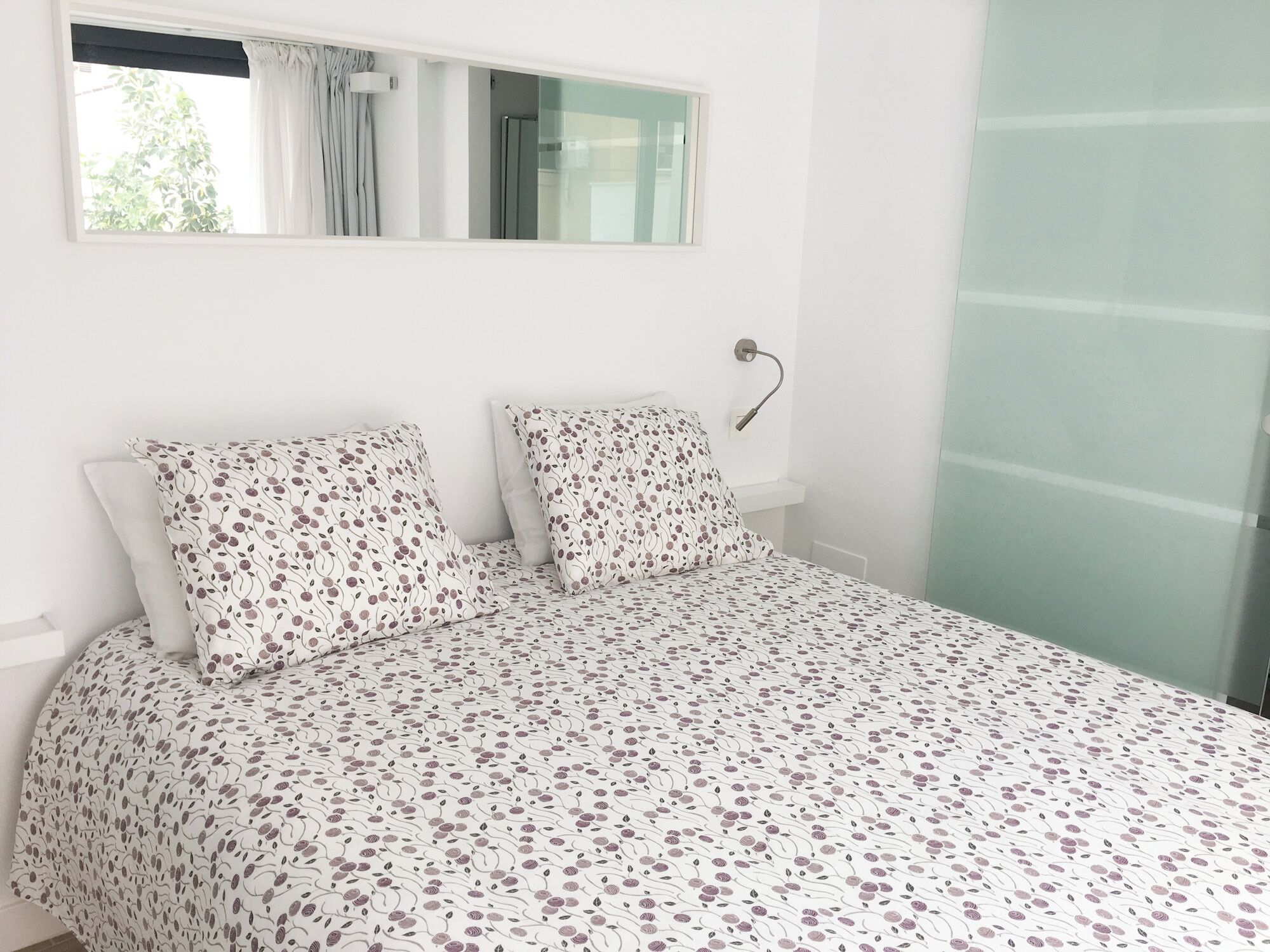 Un dormitorio con cama, dos almohadas, una mesita de noche, una ventana y un cerramiento de vidrio.