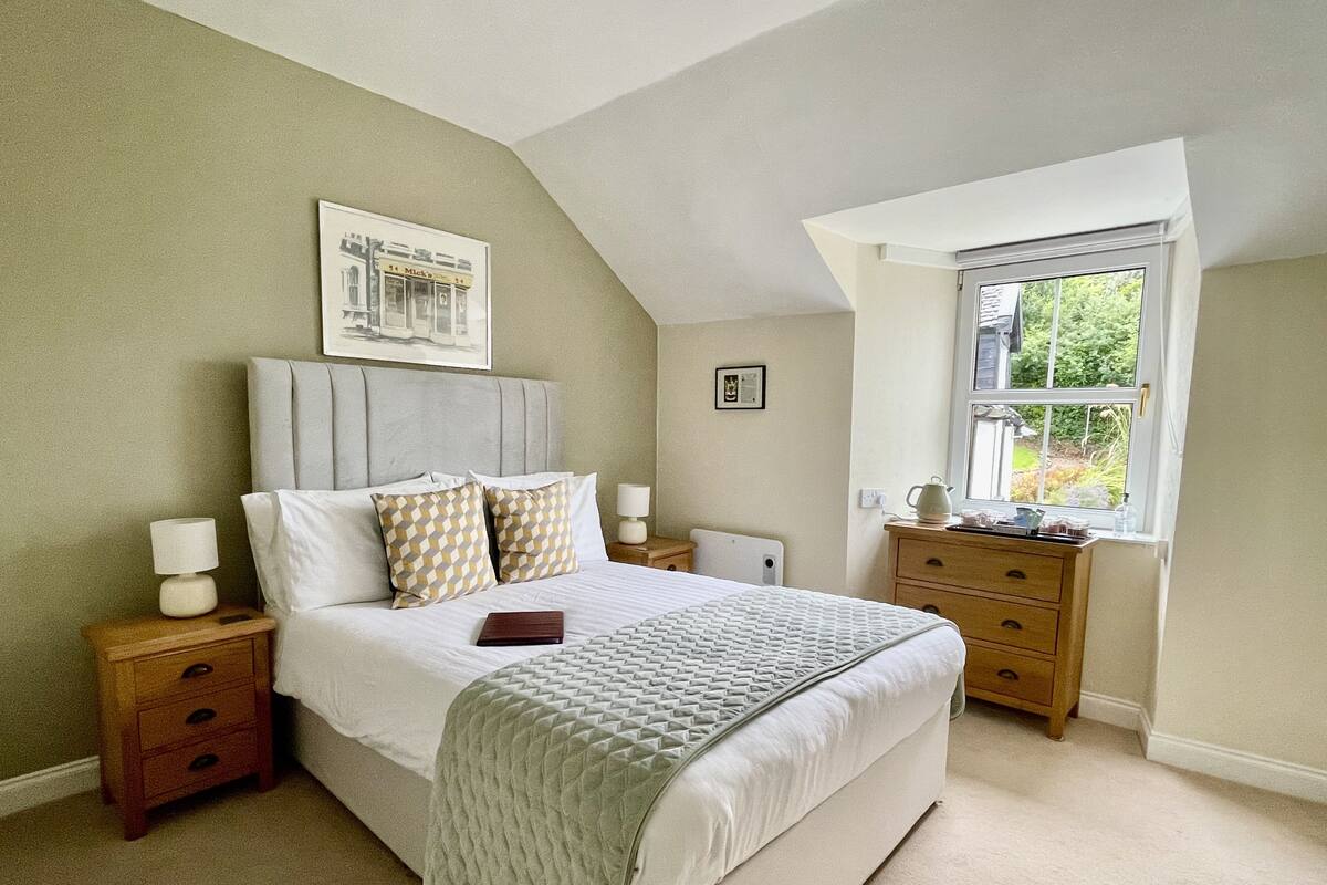 A bedroom with a large bed, a nightstand, a dresser, a window with a view, and a framed picture on the wall.