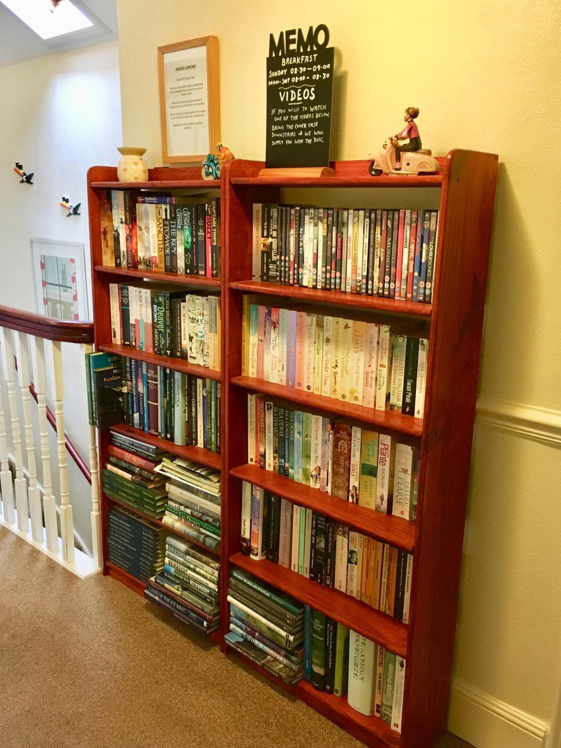 A bookshelf filled with books and DVDs, a framed sign above it, and a small figurine on top.