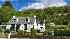 A white cottage with a stone roof, surrounded by lush greenery and a stone wall. - TwoStones (Arrochar)