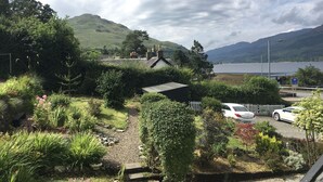 A garden with a gravel path, a white car parked nearby, and a view of a lake and mountains in the distance. - TwoStones (Arrochar)