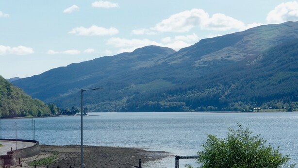 A lake with mountains in the background, a road curving around the lake, and a bridge in the foreground. - TwoStones (Arrochar)