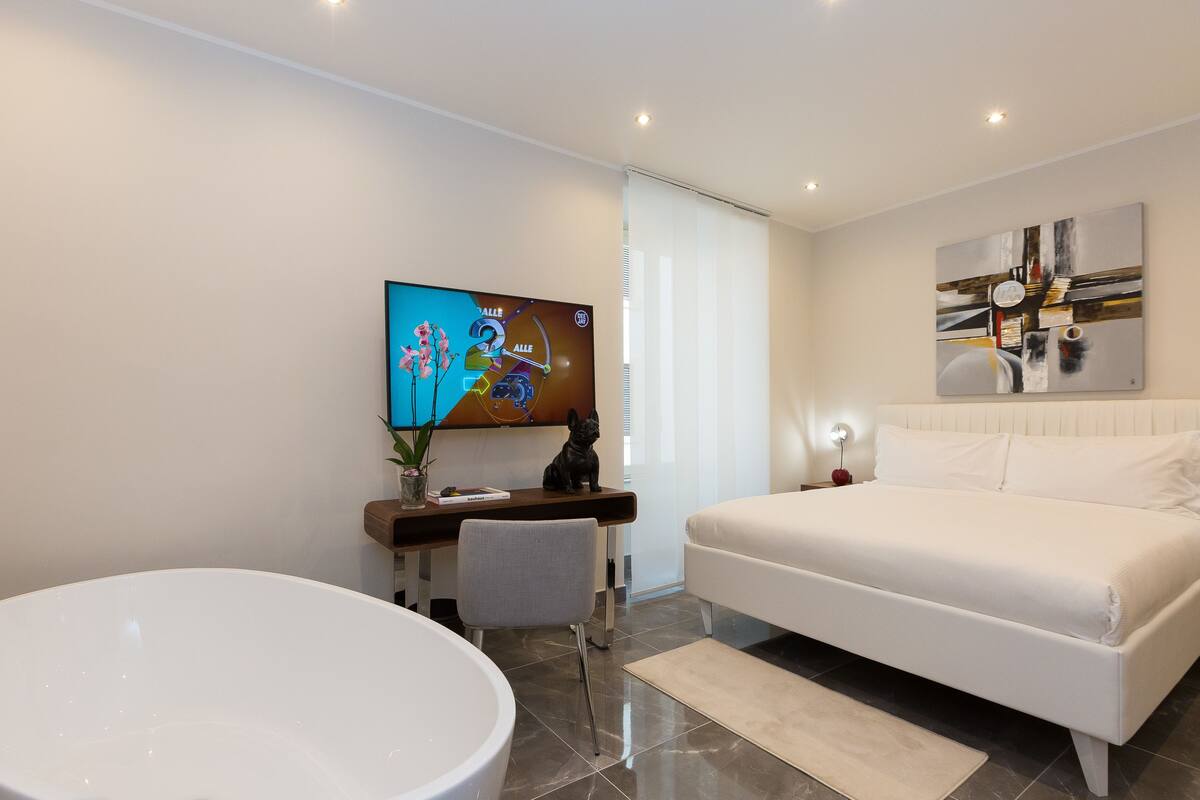 Una habitación de hotel moderna con una cama grande, un escritorio con una computadora, un televisor y un baño con un lavamanos amplio.