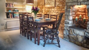 A dining area with a stone wall, wooden table, and chairs. There is a bookshelf with books and a vase with flowers. - Century House (Moreton-in-Marsh)