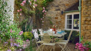 A stone building with a garden patio featuring a table and chairs, surrounded by various plants and flowers. - Century House (Moreton-in-Marsh)