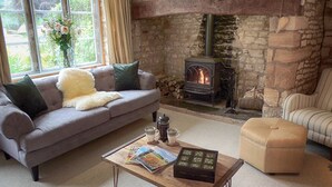A cozy living room with a stone fireplace, a grey sofa, a wooden coffee table with magazines, and a striped armchair. - Century House (Moreton-in-Marsh)