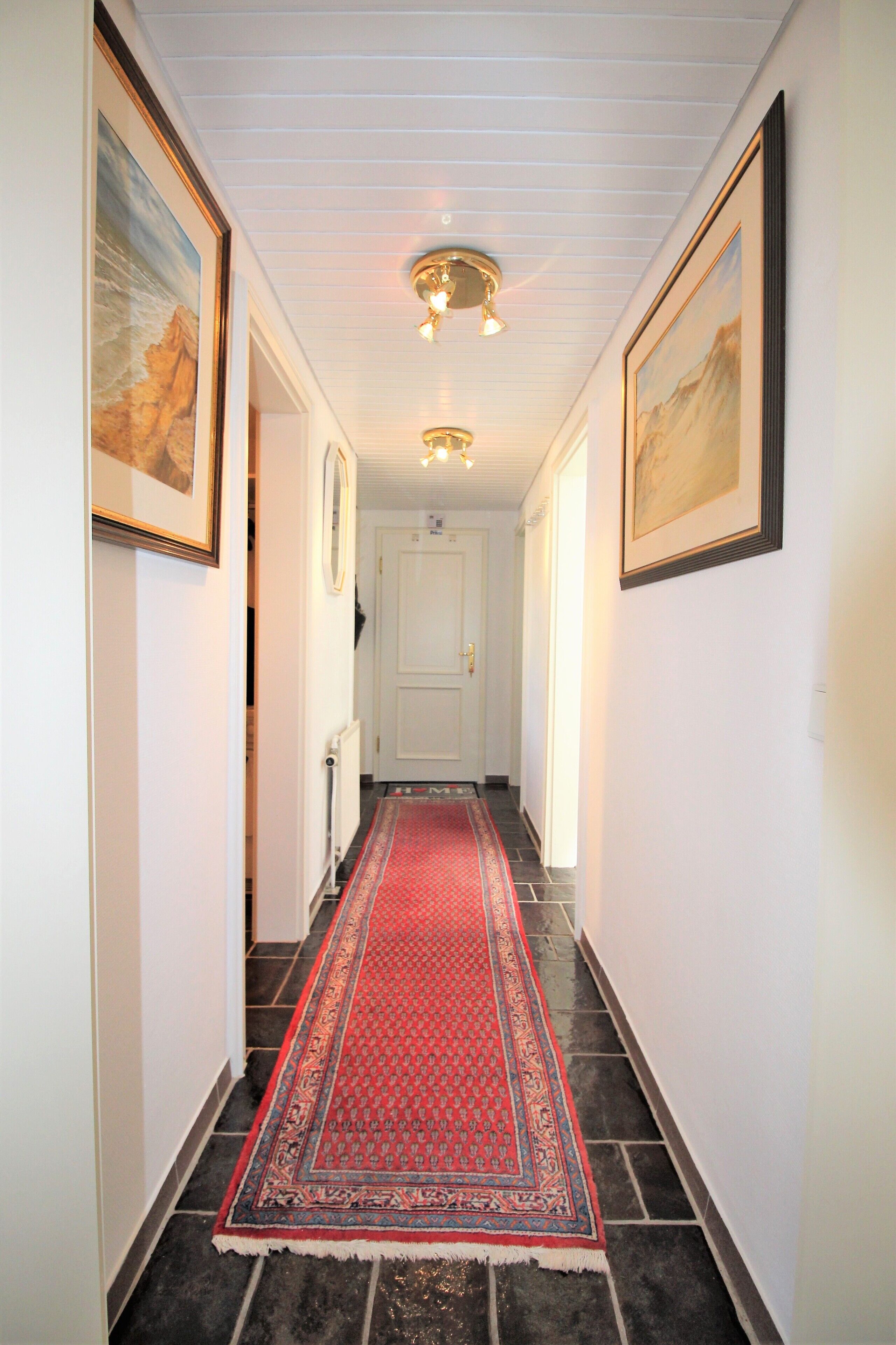 Un couloir avec un tapis rouge à motifs, des murs blancs et des œuvres d’art encadrées.
