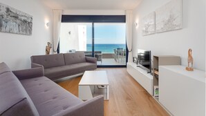 A modern living room with a sofa, a coffee table, and a TV on a white stand. There is a balcony with a view of the sea. - The Links I 2226 Golf & Sea views (La Alcaidesa)