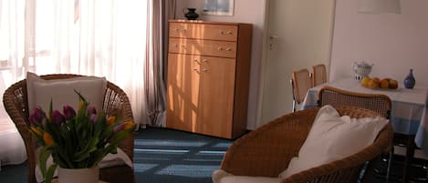 Una habitación con una silla de mimbre, una mesa con un jarrón de flores, un armario y un comedor con una mesa preparada para dos.