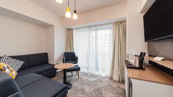 A modern living room with a sofa, armchair, and a small round table. There is a kitchen area with a countertop and a flat-screen TV mounted on the wall.