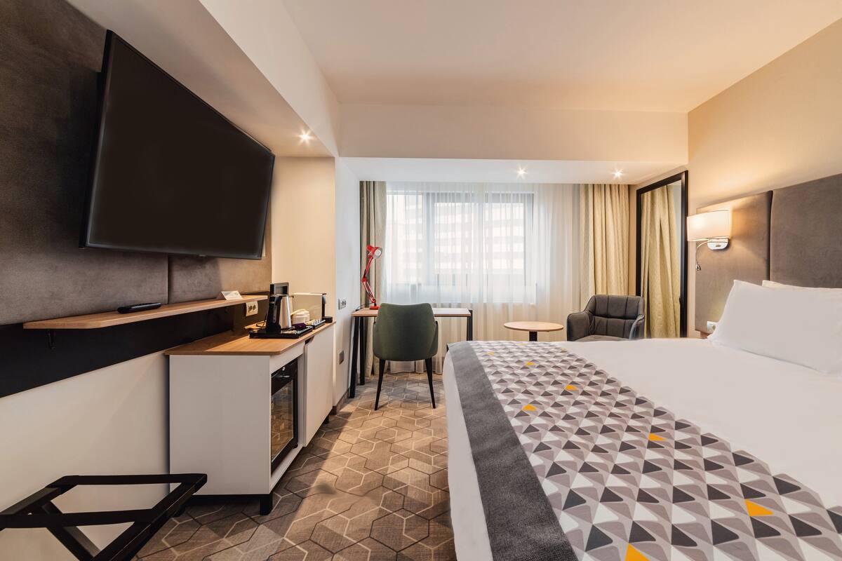 A hotel room with a bed, a desk with a chair, a television, and a window with curtains.