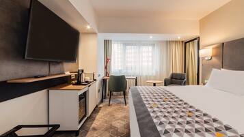 Quarto de hotel com uma cama, uma mesa de trabalho com cadeira, televisão e janela com cortinas.