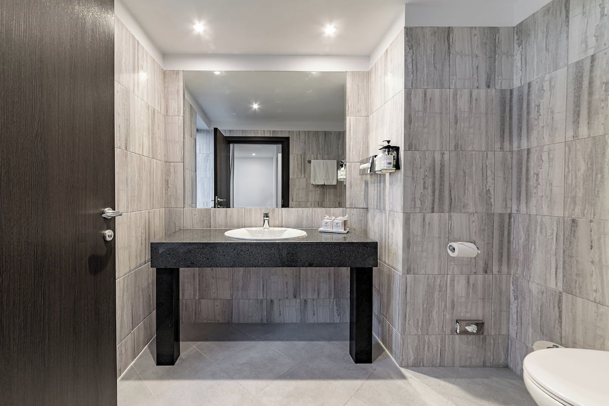A modern bathroom with a dark granite countertop, a white sink, and a large mirror above it. The walls are tiled with a light gray stone pattern, and there is a toilet to the right.