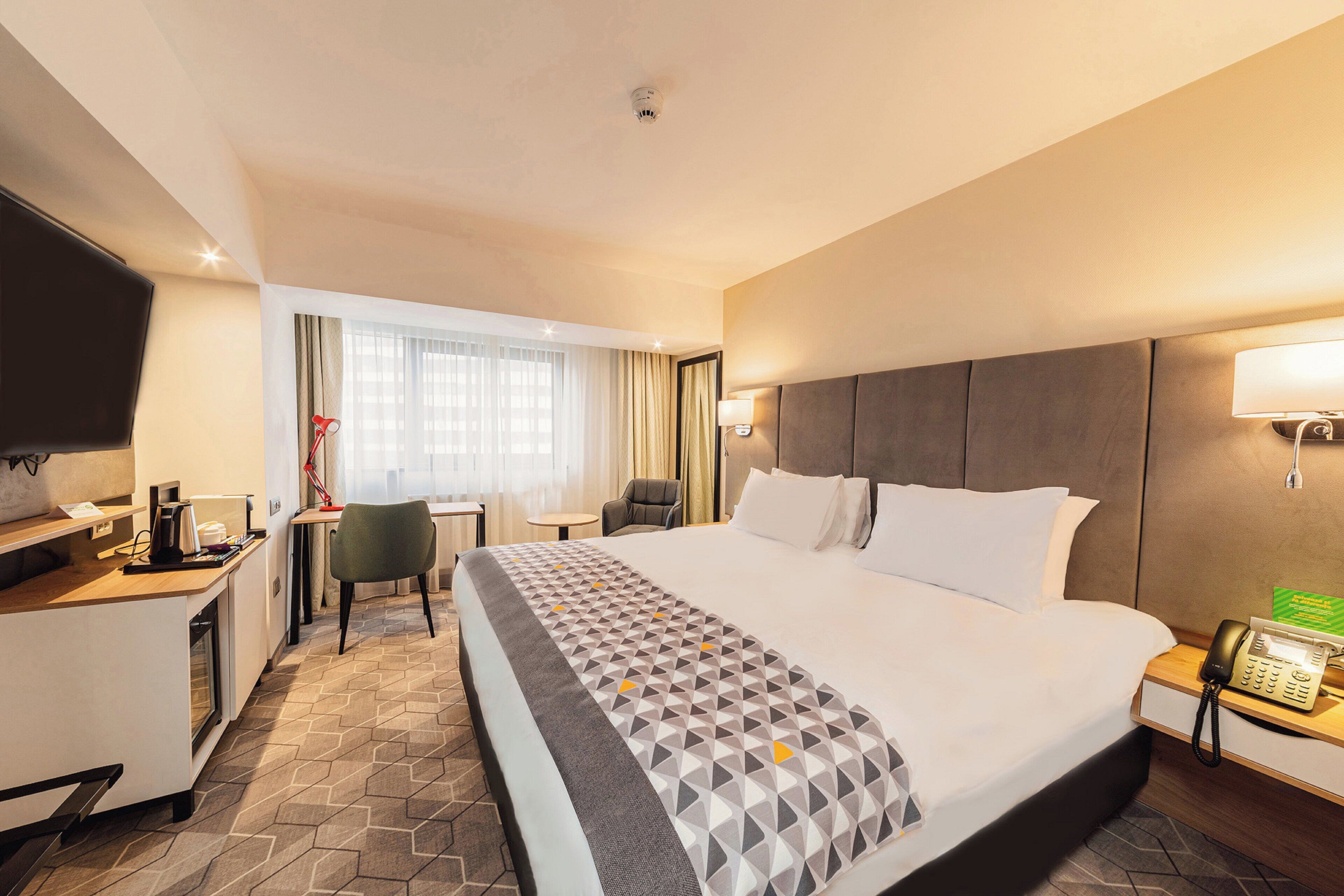 Habitación de hotel con una cama grande, un escritorio con computadora, una silla, un televisor y un teléfono.