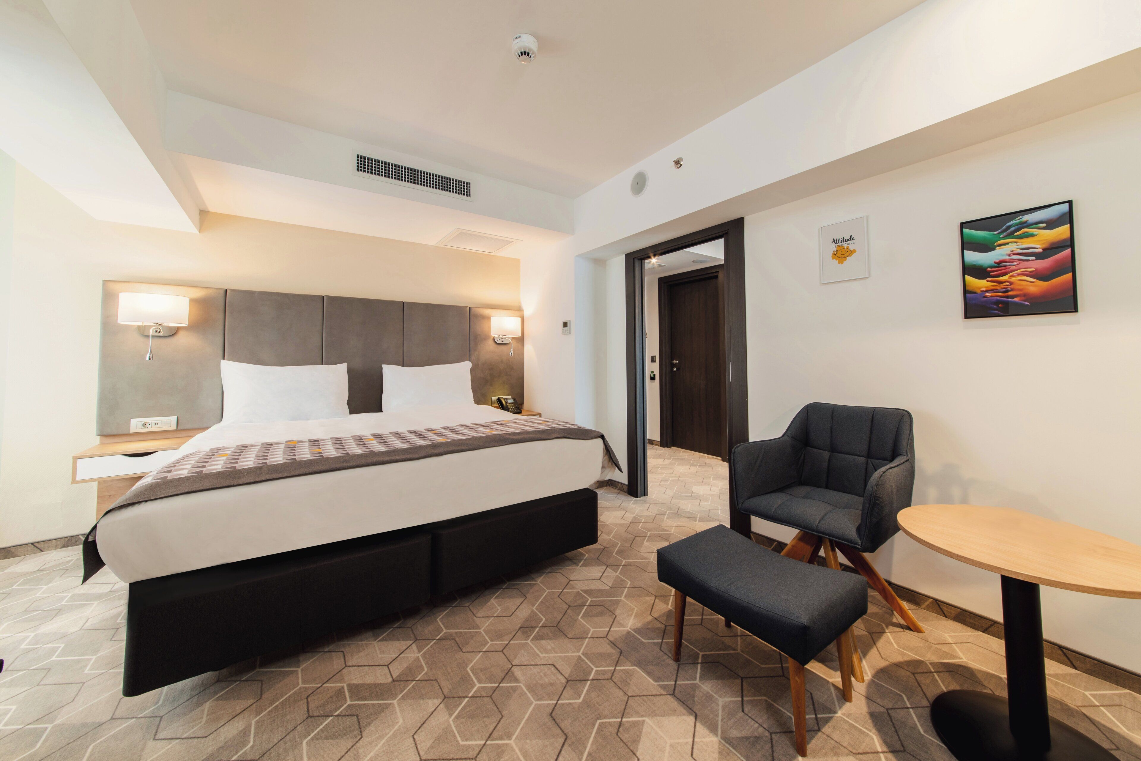 A modern hotel room with a large bed, a desk, a chair, and a small table.