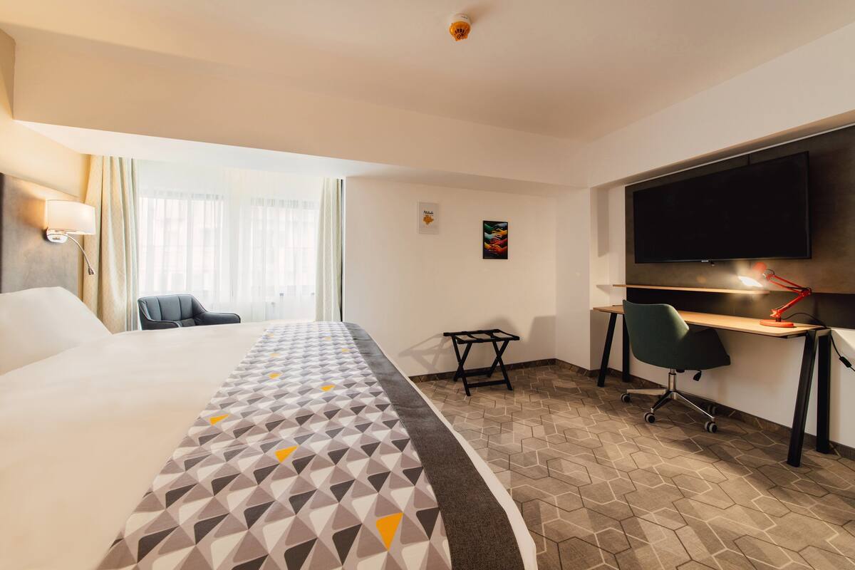 A hotel room with a large bed, a desk with a computer, a chair, and a TV mounted on the wall.