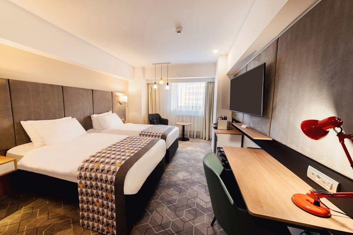 A hotel room with two beds, a desk, a TV, and a red desk lamp.