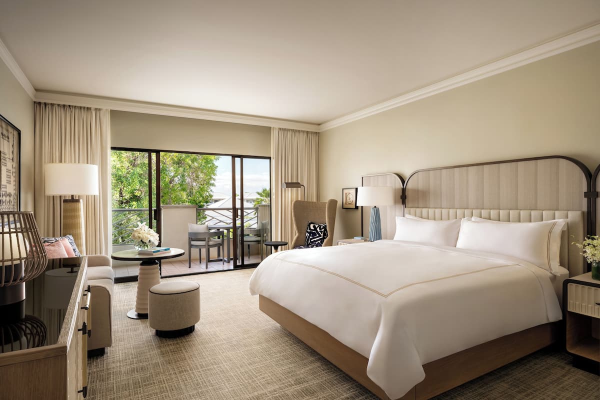 A spacious hotel room with a large bed, a sitting area with a table and chairs, and a balcony with a view of greenery.
