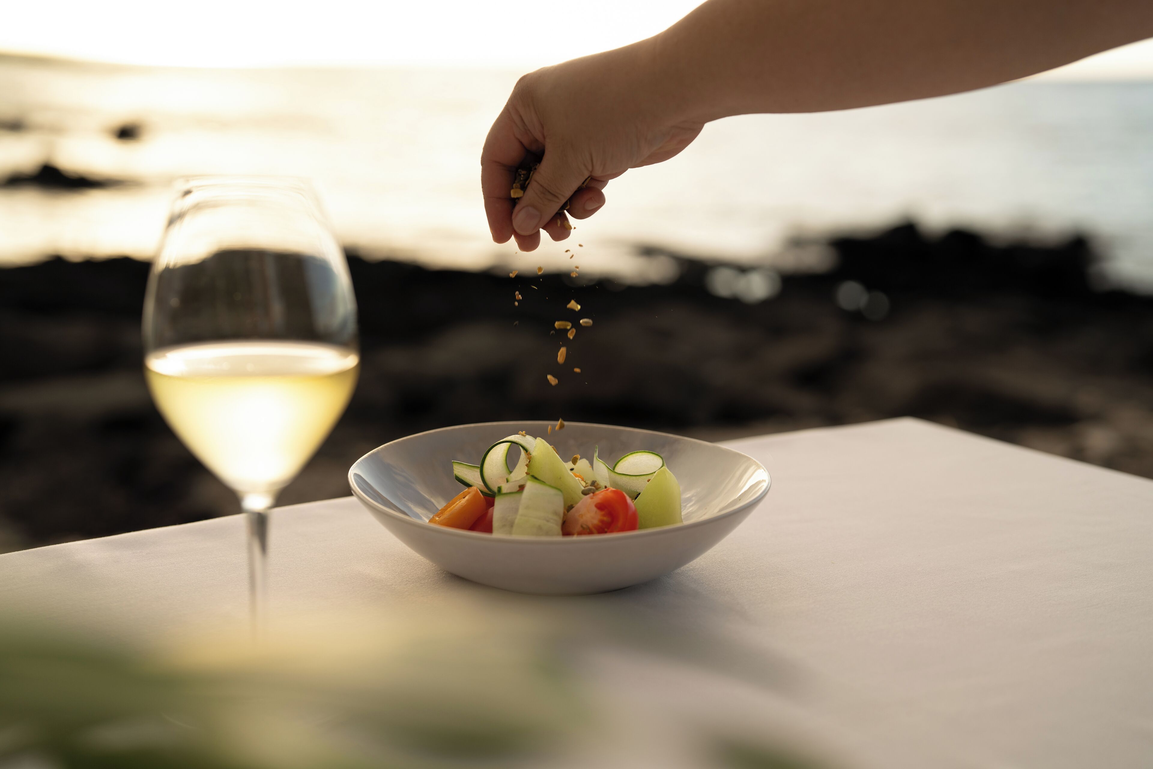 Een saladekom op een tafel met een glas witte wijn en een hand die kruiden strooit.