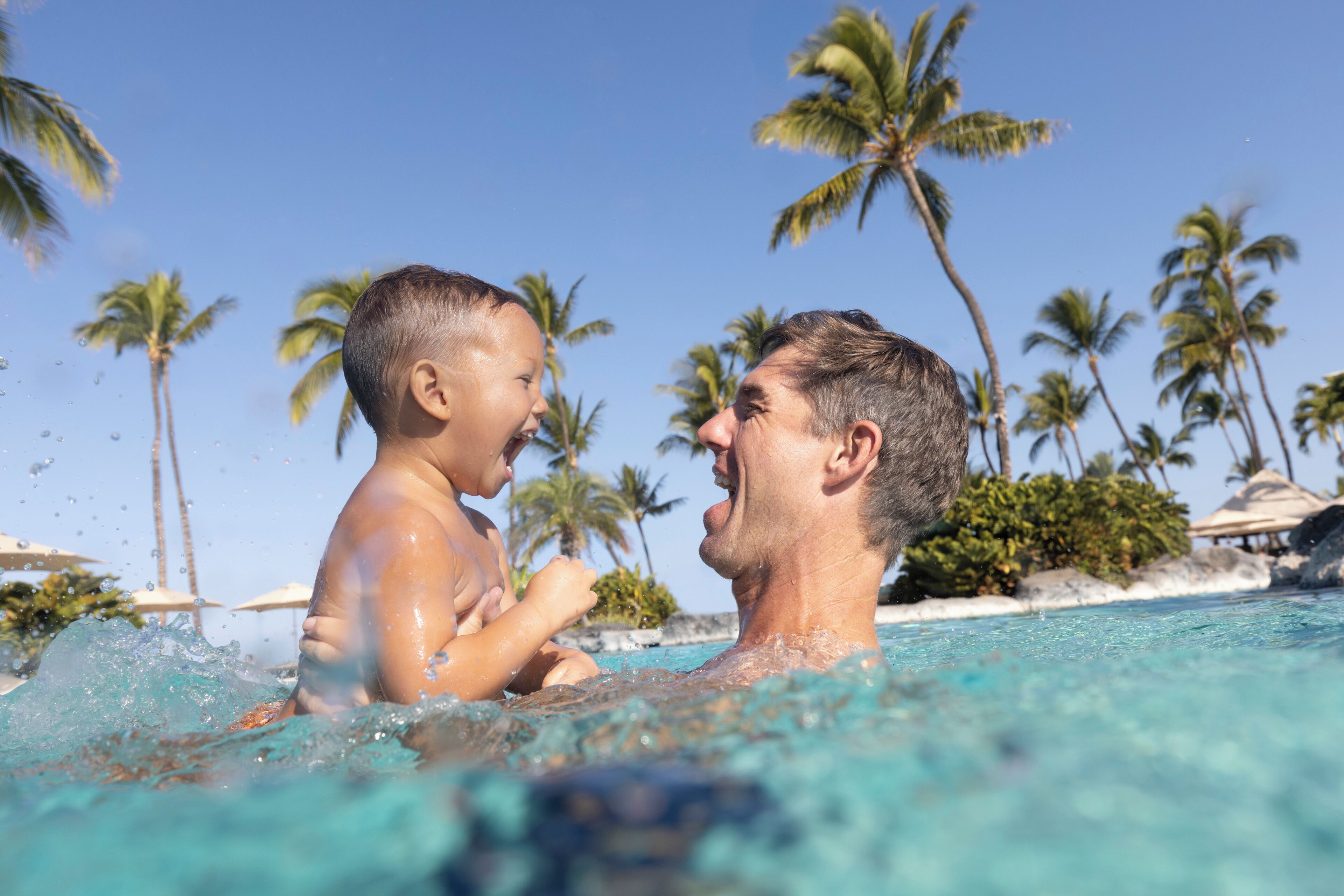 A man and a child in a swimming pool with palm trees and a clear sky in the background.