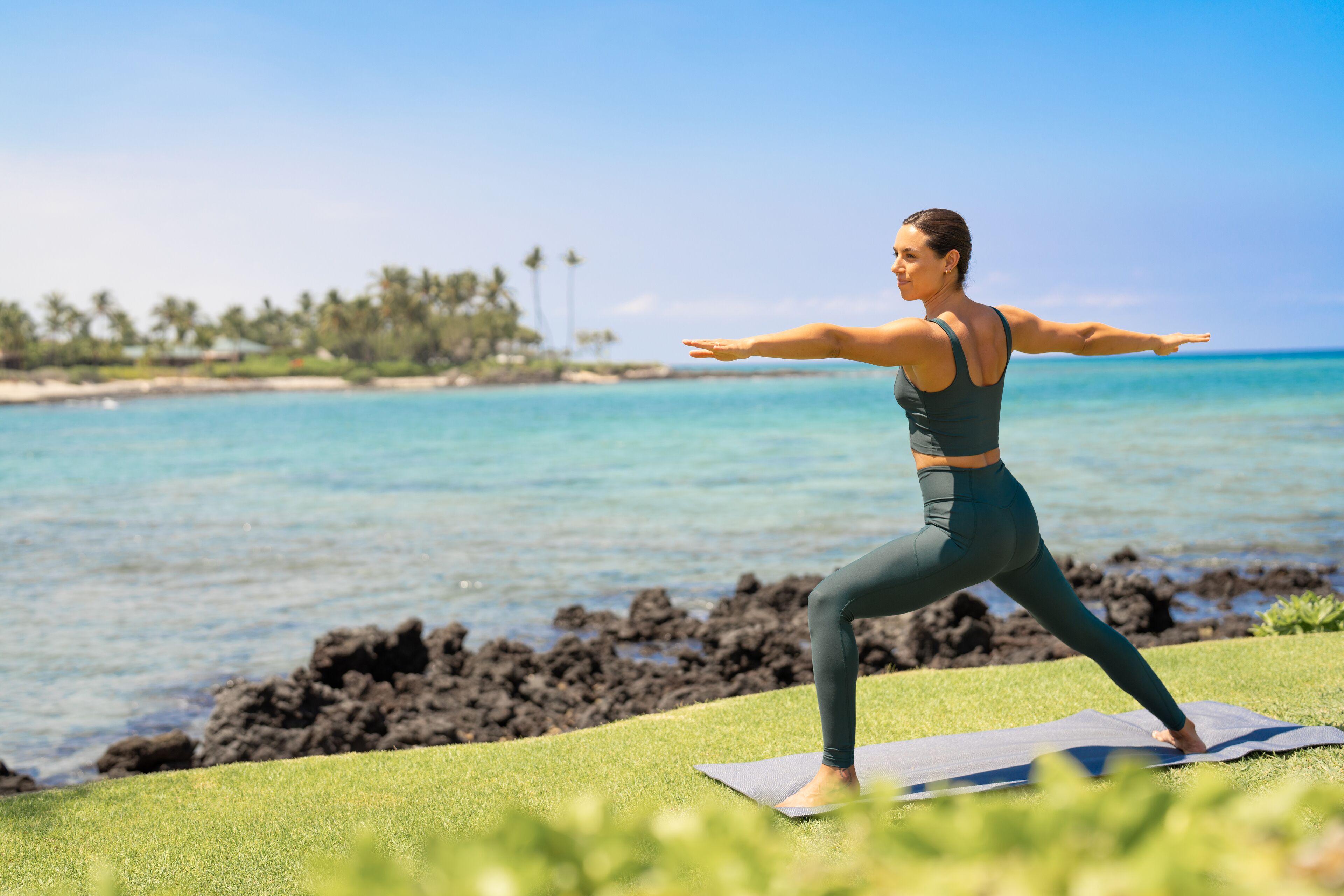 Eine Person übt Yoga auf einer Matte am Strand, mit klarem Wasser und einer entfernten Küstenlinie.