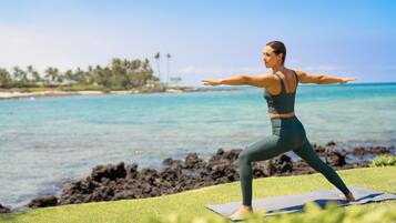 Eine Person übt Yoga auf einer Matte am Strand, mit klarem Wasser und einer entfernten Küstenlinie.