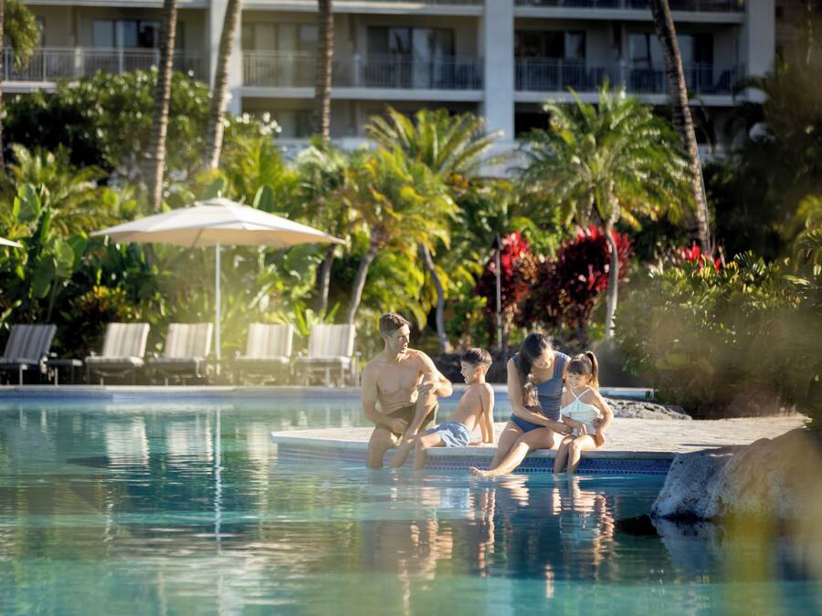 A swimming pool with lounge chairs and umbrellas, surrounded by palm trees and a hotel building.