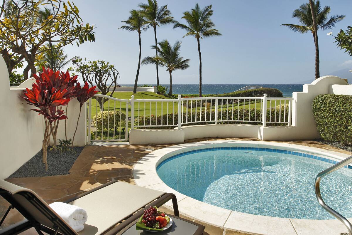 A pool area with a view of the ocean, surrounded by palm trees and a white fence.