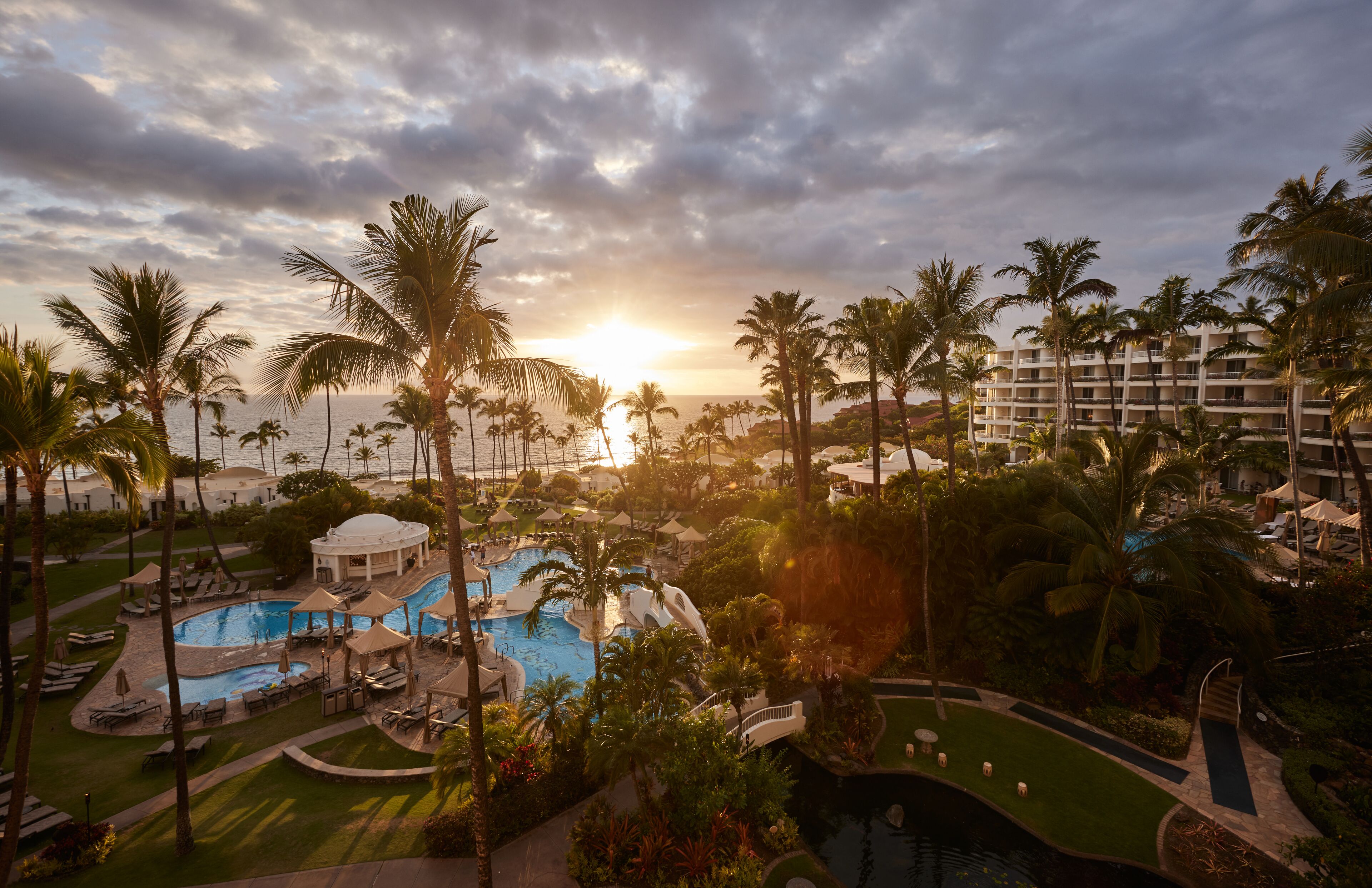 Resort com ampla área de piscina, palmeiras e um prédio com várias varandas.