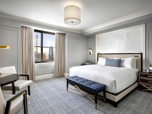 A hotel room with a large bed, a desk, a chair, a bench, a nightstand, and a window with a cityscape view.