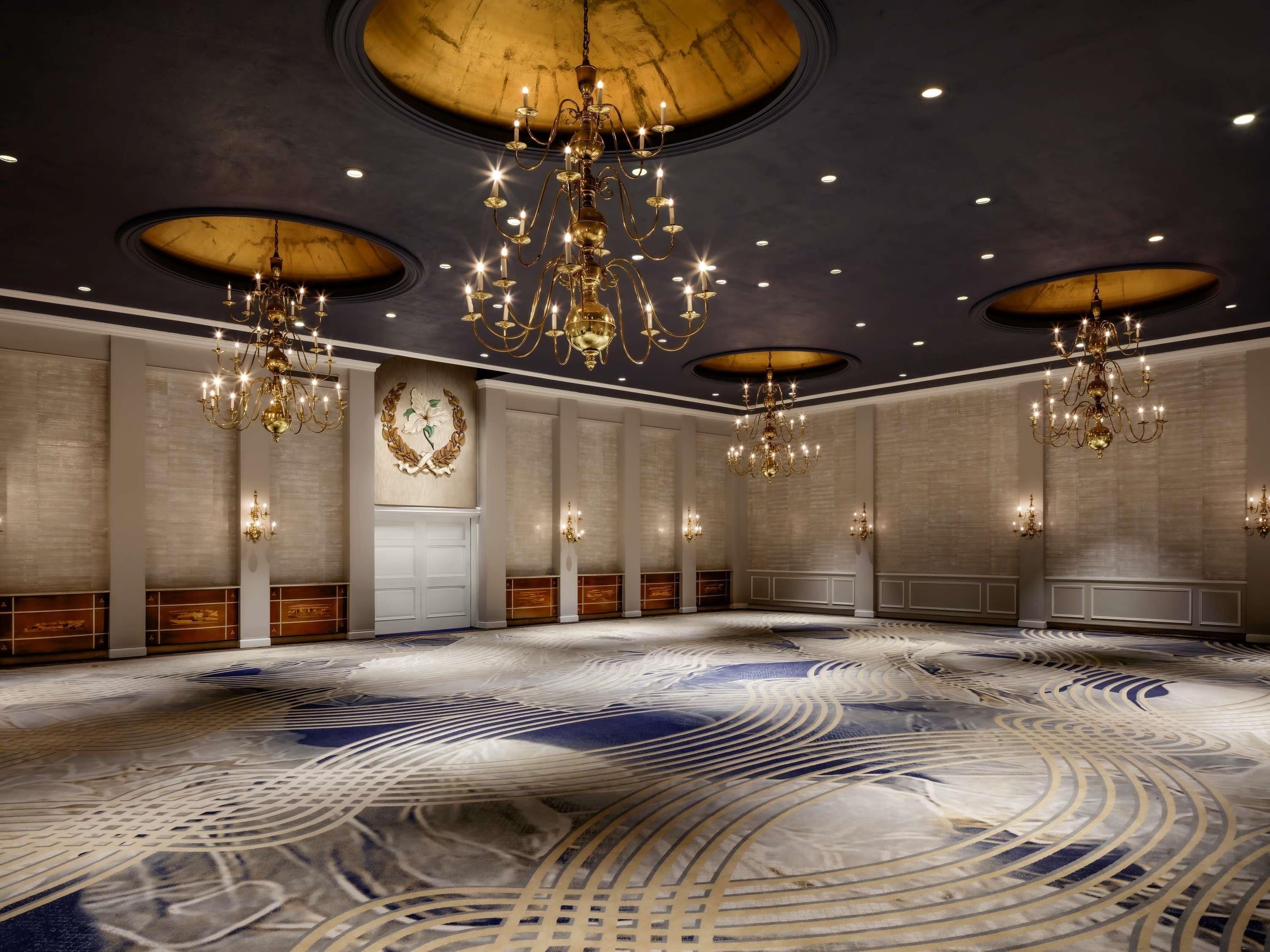 Un salón espacioso con múltiples candelabros, una alfombra con diseño y una gran pared con un emblema decorativo.