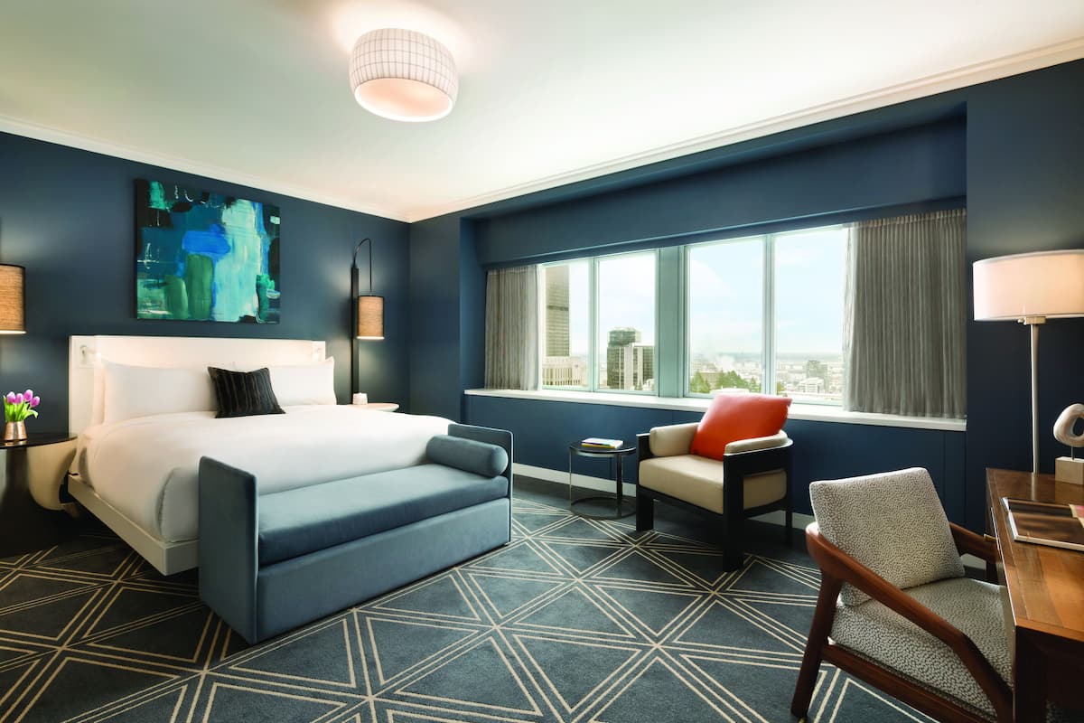 A modern hotel room with a large bed, a seating area, a view of the city, and a geometric patterned carpet.