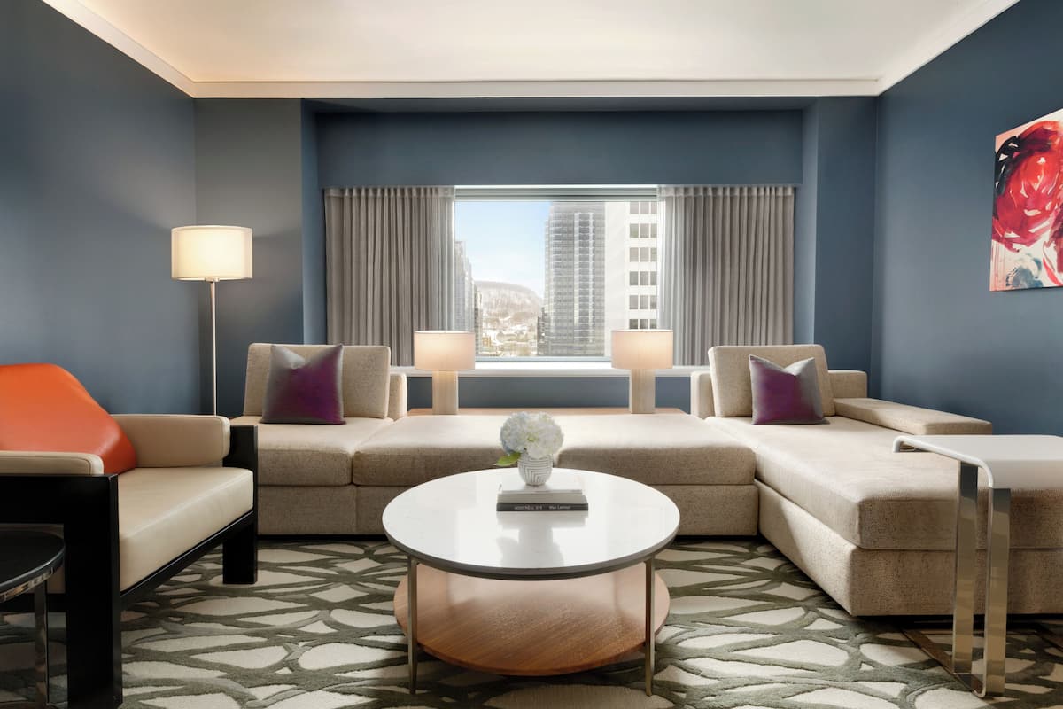A modern hotel room with a sectional sofa, a round coffee table, and a view of the cityscape through large windows.