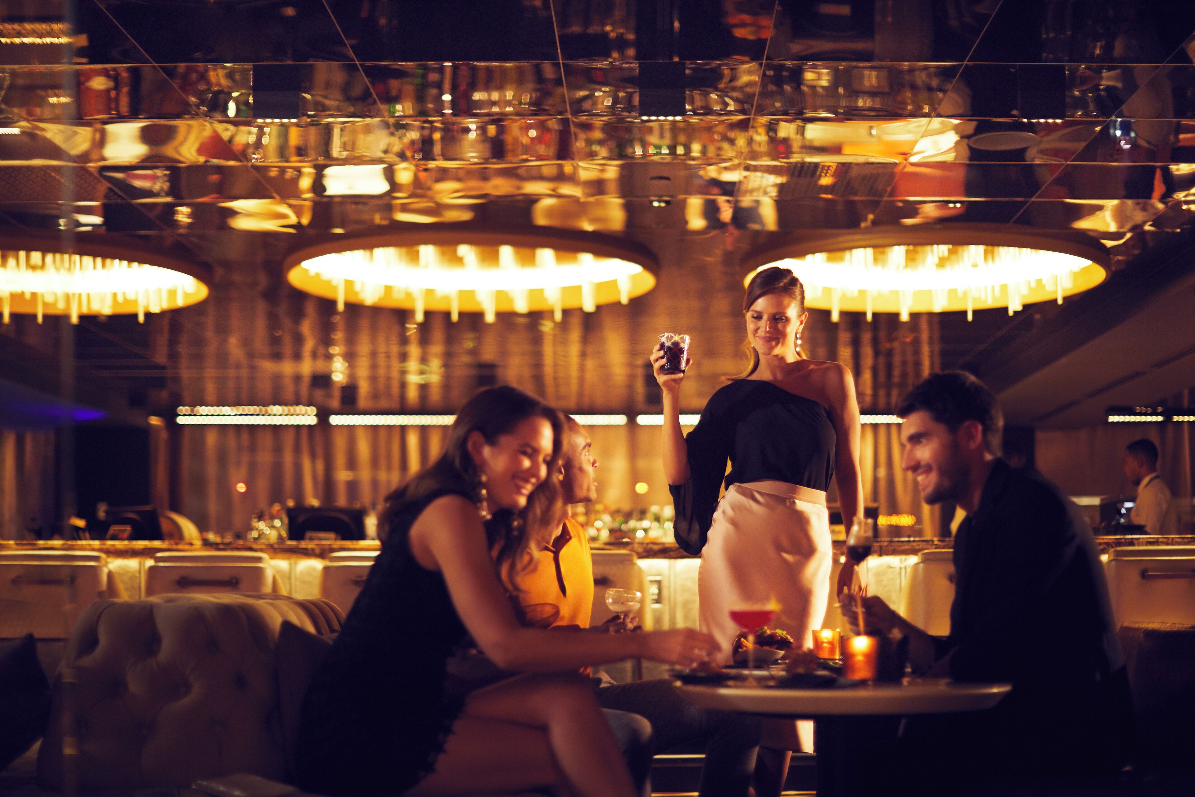 A dimly lit bar with multiple hanging lamps, a bartender, and patrons seated at tables.