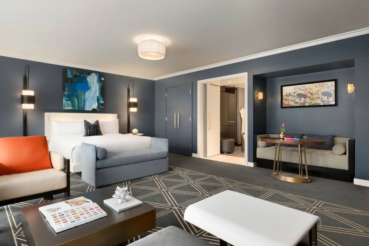 A modern hotel room with a bed, a sofa, a small table, and a desk. There is a door leading to a bathroom and a hallway.