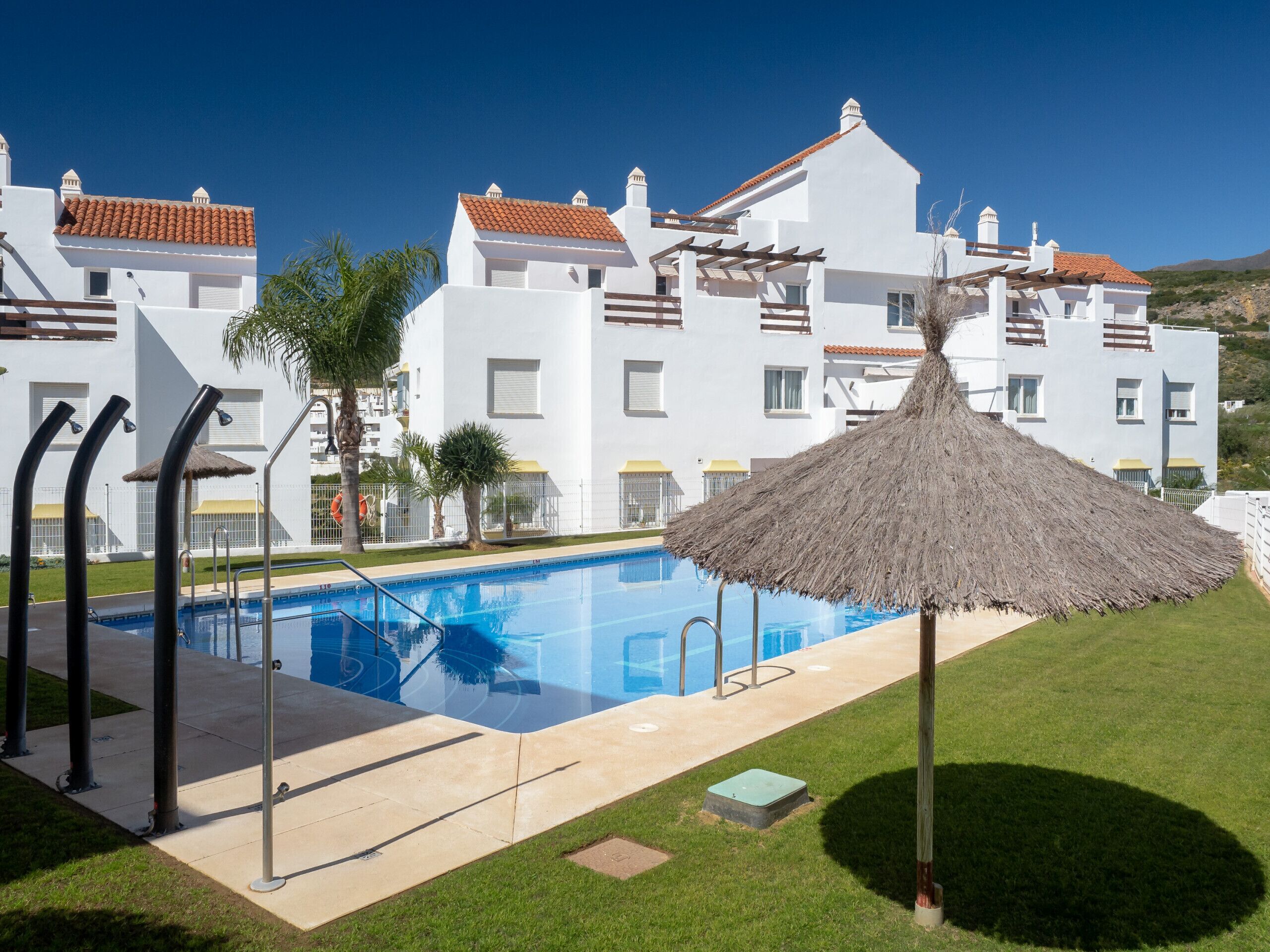 Une piscine agrémentée d’un parasol en chaume, entourée d’une pelouse bien entretenue et, au fond, d’un bâtiment blanc dont le toit est recouvert de tuiles rouges.