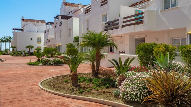 Ein modernes Hotel mit weißen Fassaden, mehreren Balkonen und einem angelegten Garten mit Palmen und Blumen.