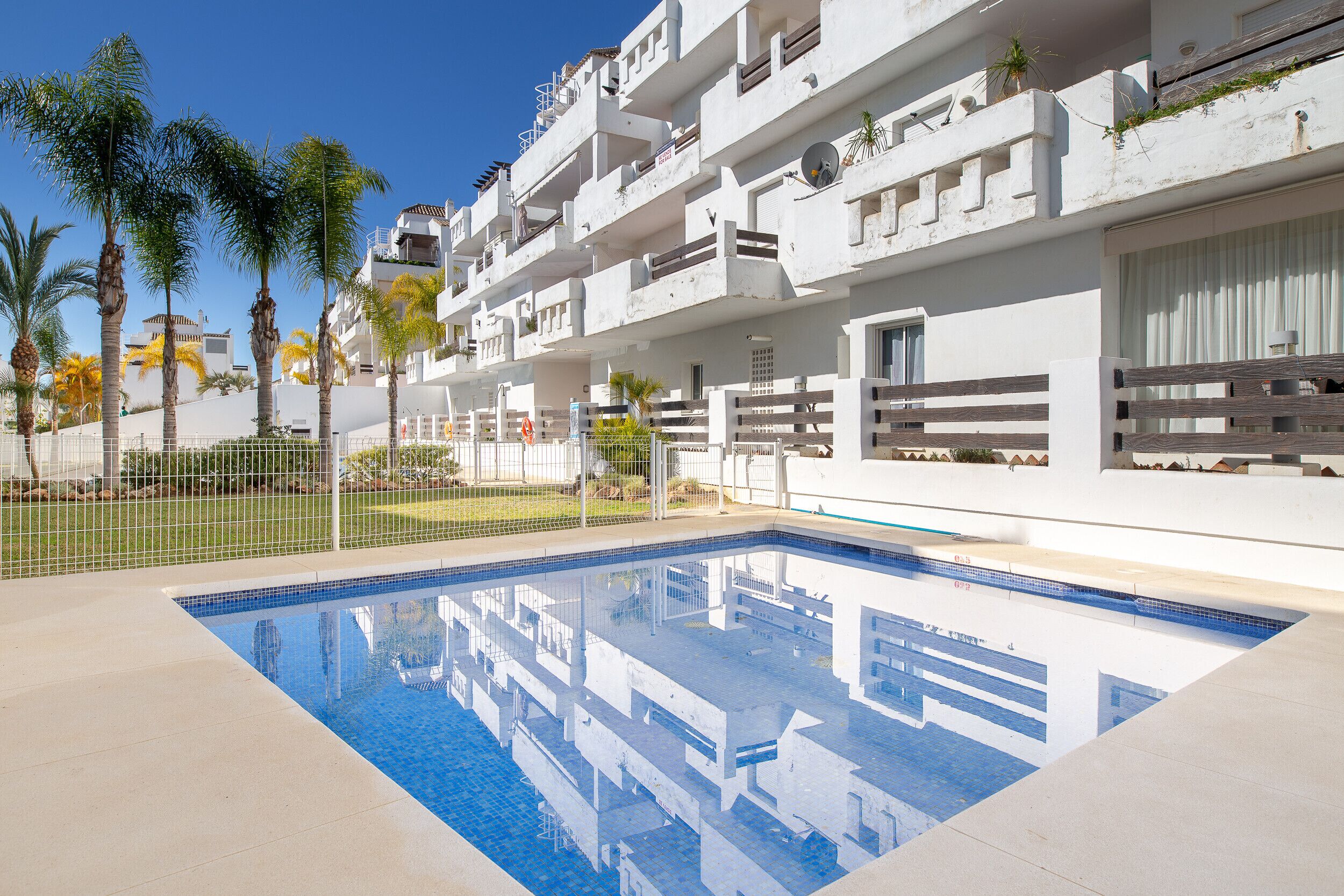 A swimming pool with a clear blue water surface, surrounded by a white building with balconies and a fenced area with palm trees.